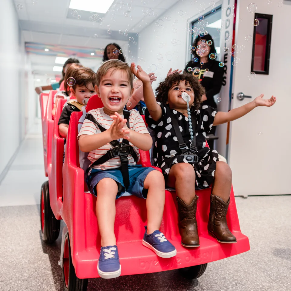 Children smiling and reaching toward bubbles in a red wagon.